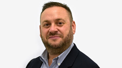 A man with short brown hair and a full beard, wearing a dark navy blazer over a light blue button-down shirt