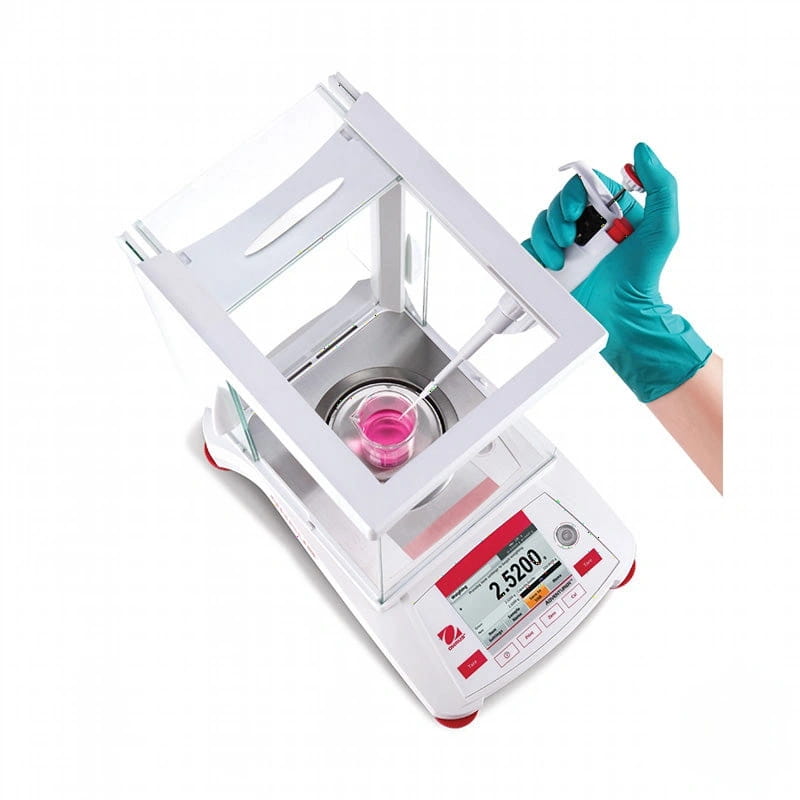 White analytical balance with red accents and digital display showing 2.5200 grams, featuring a transparent draft shield over the weighing pan containing pink liquid being dispensed into it by a pipette held in gloved hands