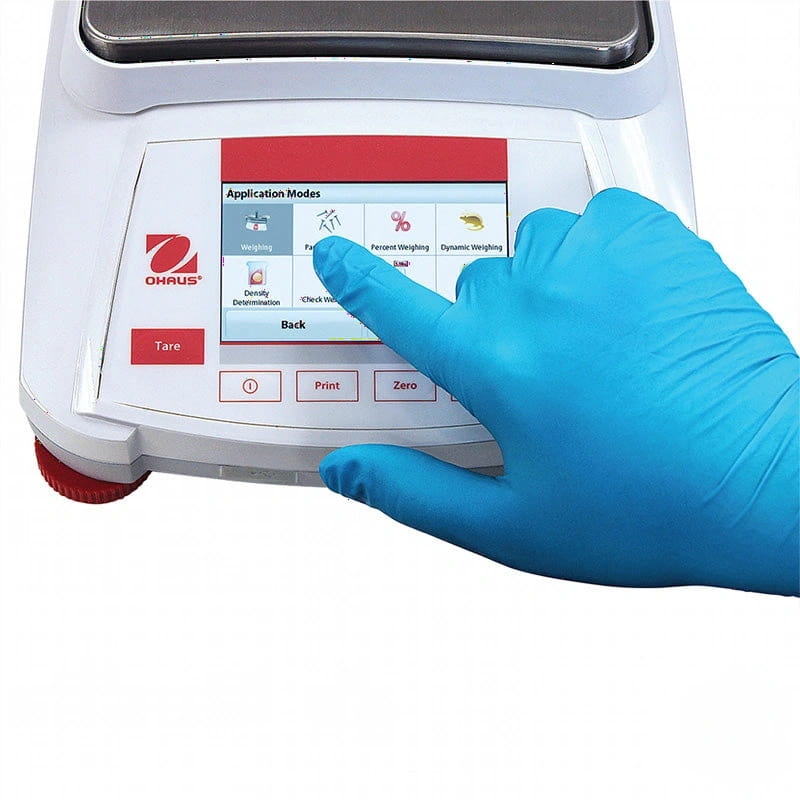 White ohaus digital laboratory scale with a touchscreen displaying application modes and a blue-gloved hand interacting with its controls