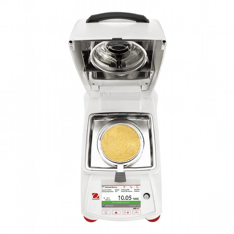 White analytical balance with a digital display showing 10.05 grams, featuring a transparent sample pan containing yellow powder and an open upper chamber revealing internal components